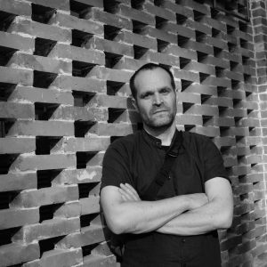 Seth wears a dark button up shirt and leans against a brick wall with folded arms looking toward the camera.